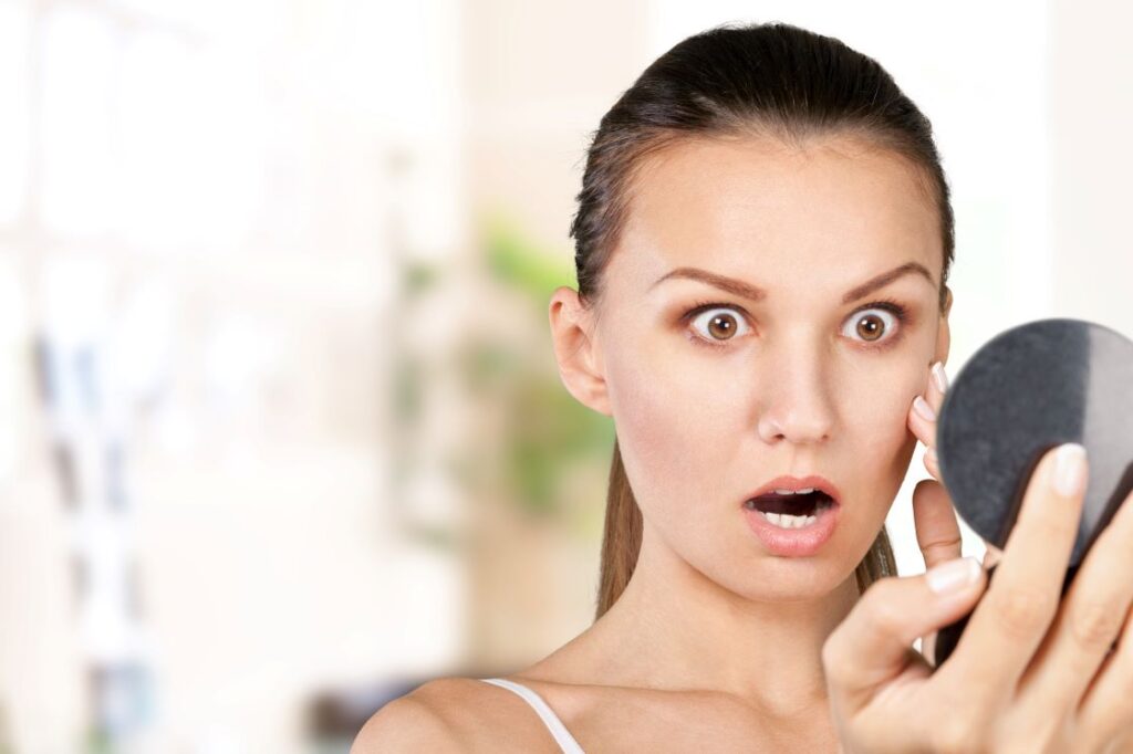 A woman looking shocked at her appearance in a mirror.