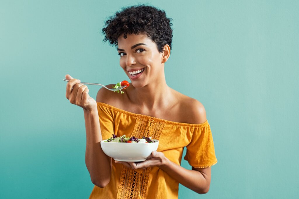 Woman with dark curly hair eating salad smiling in front of teal background